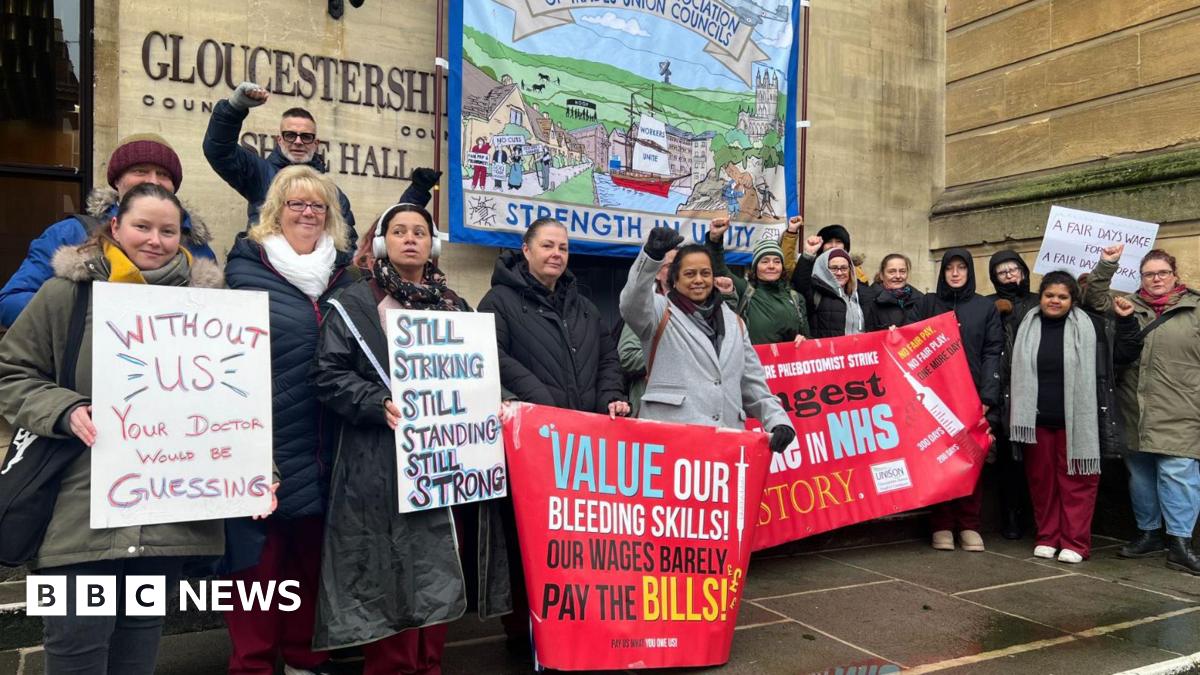 A group of striking NHS workers - perhaps around 20 people - stand in front of Gloucestershire's Shire Hall holding placards with slogans such as "Without us, your doctor would be guessing" and "Still striking, still standing, still strong".