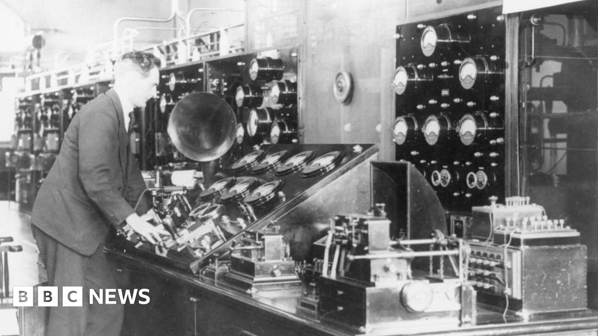 A male operator stands at the main control table containing all the main control instruments including many dials and switches.  It is a black and white image and he is wearing a dark suit and tie.