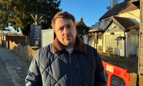 A man in a dark coat is looking at the camera. He is in front of a pub site, which has the name The Hare & Sounds on a board and on the outside of the building. 