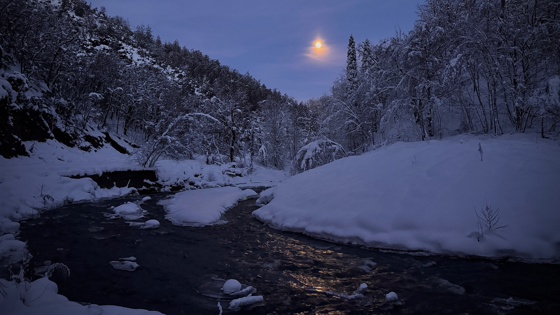 An orange moon is pictured glowing above a snowy hillside covered in trees at night, as a river flows freely in the foreground, reflecting its light.