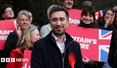 Damien Egan wearing a red rosette, stands in front of a group of people, some are holding placards reading: "Britain's Future"