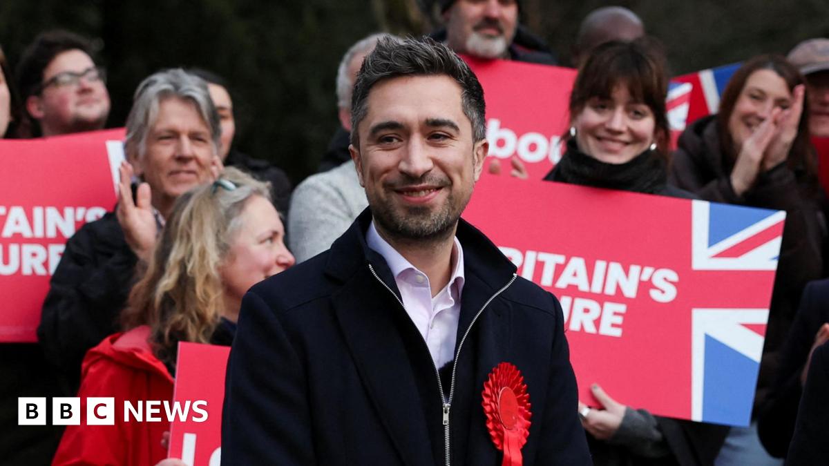 Damien Egan wearing a red rosette, stands in front of a group of people, some are holding placards reading: "Britain's Future"