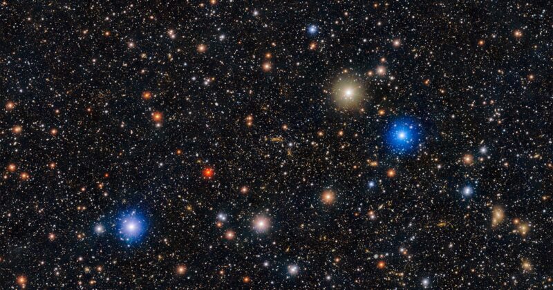 A dense field of stars and galaxies in deep space, featuring various bright blue, white, yellow, and red points of light scattered across a dark background.