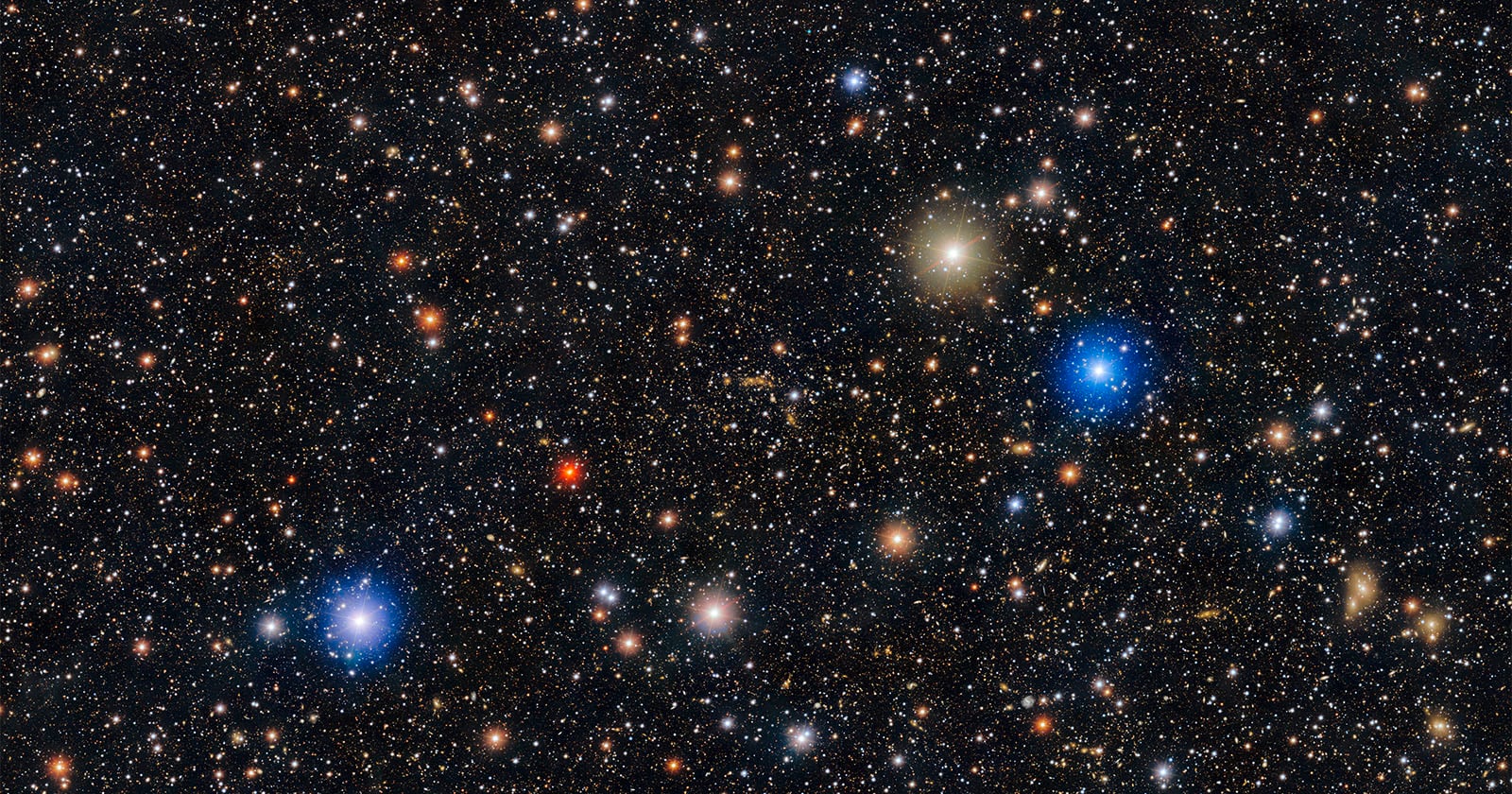 A dense field of stars and galaxies in deep space, featuring various bright blue, white, yellow, and red points of light scattered across a dark background.