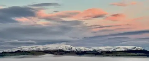 David May Snow‑covered hills stretch across the horizon beneath a sky with pink and blue clouds. A layer of mist lies in the valley below, while the foreground shows gentle green slopes partly obscured by fog.