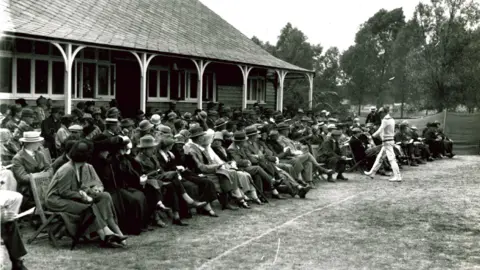 Essex County Cricket Club A black and white image from Essex against Oxford University match. Several rows of people form the crowd in front of a clubhouse. They are dressed smartly with hats on. A cricketer wearing whites walks past them.