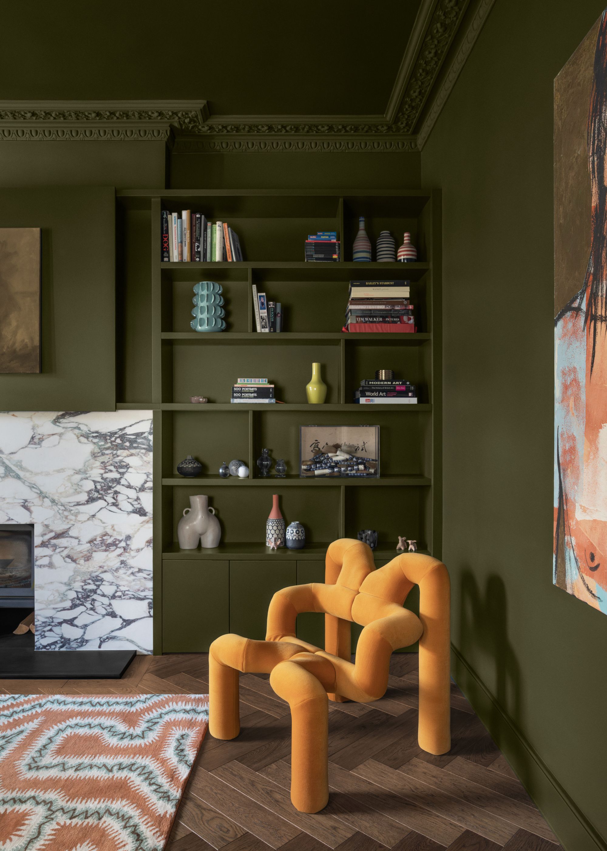 A living room with dark mossy green color-drenched walls, a marble fireplace, and a bright yellow sculptural accent chair.