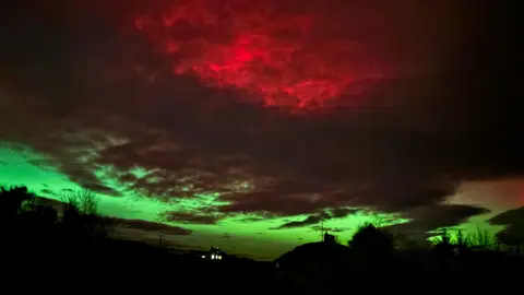 BBC Weather Watchers/Jane O Dark clouds illuminated from below by green aurora contrast with vivid red light glowing through the upper cloud layer over a rural landscape.