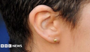 A side view of a woman's ear, the lobe of which is pierced with a gold stud. She has short dark hair.