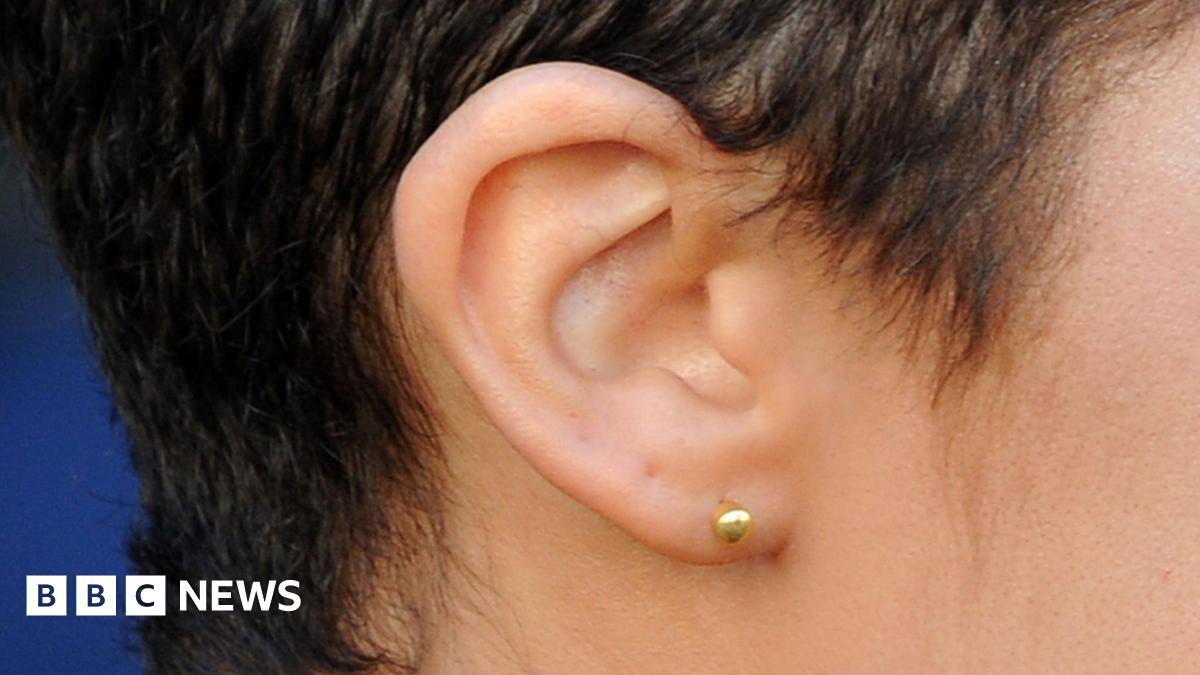 A side view of a woman's ear, the lobe of which is pierced with a gold stud. She has short dark hair.