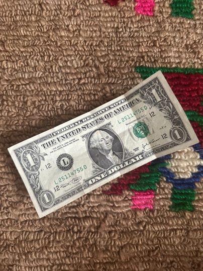 A US $1 note is seen photographed on a beige, green and red carpeted surface.