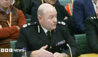 A bald man in a formal police uniform is sitting in a chair with a small microphone in front of him. There are other people around him