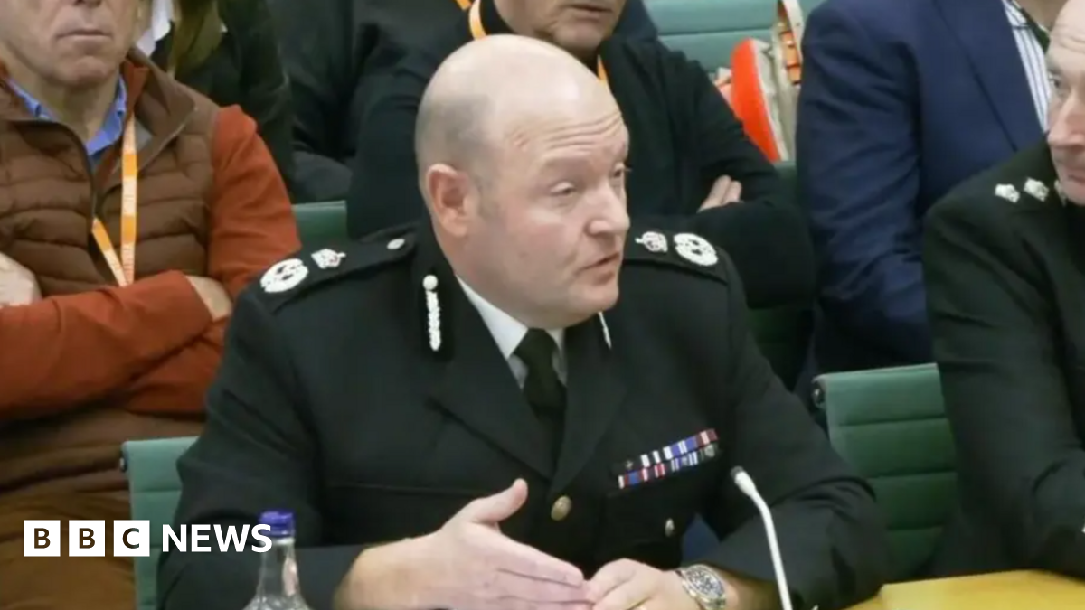 A bald man in a formal police uniform is sitting in a chair with a small microphone in front of him. There are other people around him