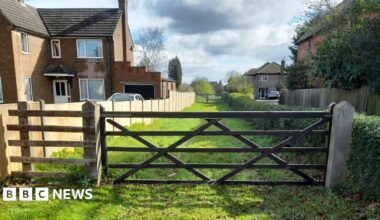 Access to former cathedral quarry site, Riseholme Road, Lincoln. The picture shows a wooden gate across a narrow stretch of grass, with houses either side.
