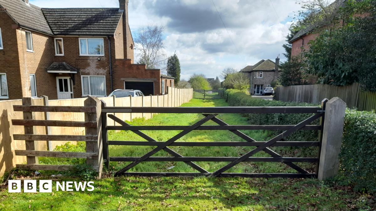 Access to former cathedral quarry site, Riseholme Road, Lincoln. The picture shows a wooden gate across a narrow stretch of grass, with houses either side.