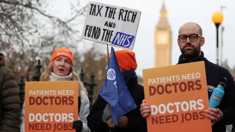 Reuters Two resident doctors stand on the picket line in London. One is a bald man with an NHS flask and black plastic rimmed glasses. He is holding a "patients need doctors, doctors need jobs" sign. A female resident doctor stands beside him and is holding the same orange poster. There is a third person with an orange beanie hat on standing behind a flag in the centre of the picture with a plaquard saying "tax the rich, fund the NHS"