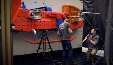 Two men in dark shirts work on a red car-sized rotorcraft protype in a testing chamber.