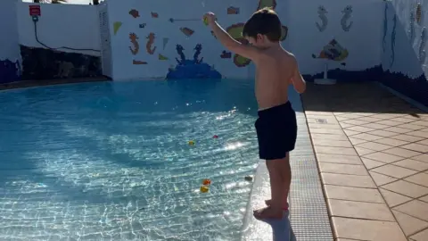 Tammy McDaid A little boy is standing by the side of a swimming pool. He appears to be dropping toys into the water. A mural featuring seahorses can be seen in the background.