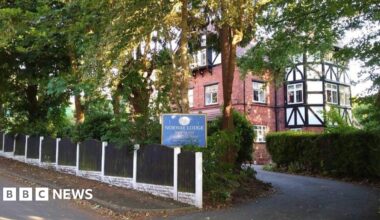 A tree and hedge lined drive way leading to a brick building with Victorian style white fascia lined with black wooden beams. A blue sign marks the entrance with 'Norway Lodge' in white letters.