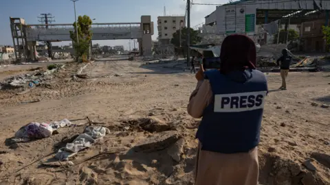 EPA/Shutterstock A journalist wearing a protective vest with the word "PRESS" across the back stands on a road strewn with debris in Gaza