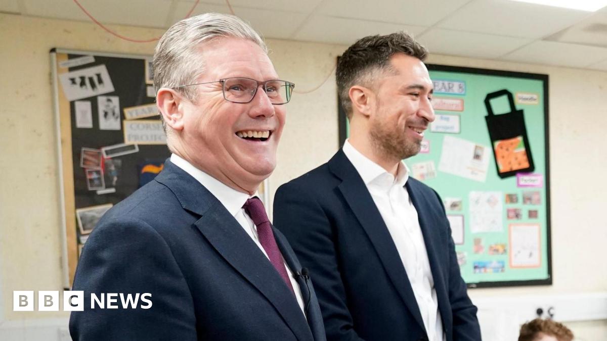 Sir Keir Starmer and Damian Egan stand together in a classroom where children are sitting at desks. Both me are wearing suits and the children are in school uniform.
