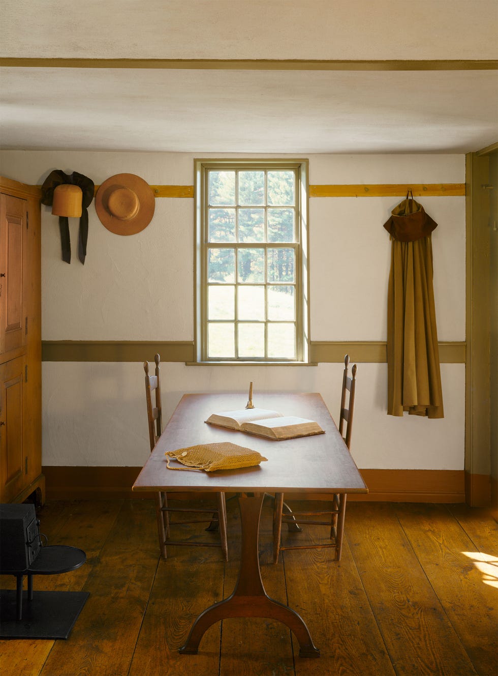 simple shaker room with rustic wood pedestal table with simple slat back chairs and a window and a wall with pegs holding hats and a caps