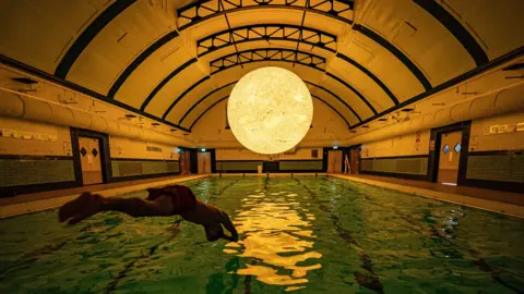 PA Media A silhouette of a swimmer diving into an indoor pool. There is a large sculpture modelled off of the sun suspended above the pool. It is reflecting in the water.