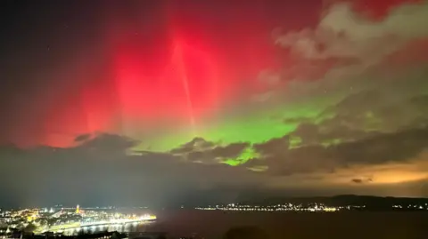 BBC Weather Watchers/RockDodger A bright red and green aurora glows above a town and coastline, with city lights reflecting off the water beneath low clouds.
