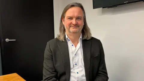 BBC Paul Gorman pictured wearing a white button down shirt and black blazer. He has shoulder-length brown hair and stubble. He is smiling at the camera without teeth.