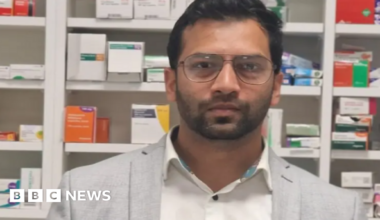 A man with a very short beard is wearing glasses, a white shirt and a grey blazer. He is standing in front of row of medication boxes.