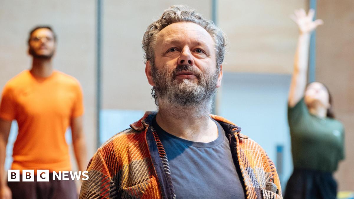 Michael Sheen in a checked shirt and T-shirt in the foreground, with other actors at the back of the rehearsal room