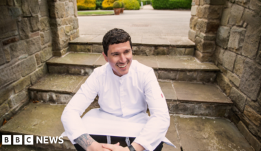 Millie Bobby Brown and David Harbour in character as Eleven and Jim Hopper. Eleven wears red shorts over grey leggings and a grey jumper. Jim Hopper wears a black leather jacket, a black beanie hat and dark trousers.They are speaking to director Frank Darabont on the set of Stranger Things. There is a vintage advertisement posted in the background with debrief on the ground.