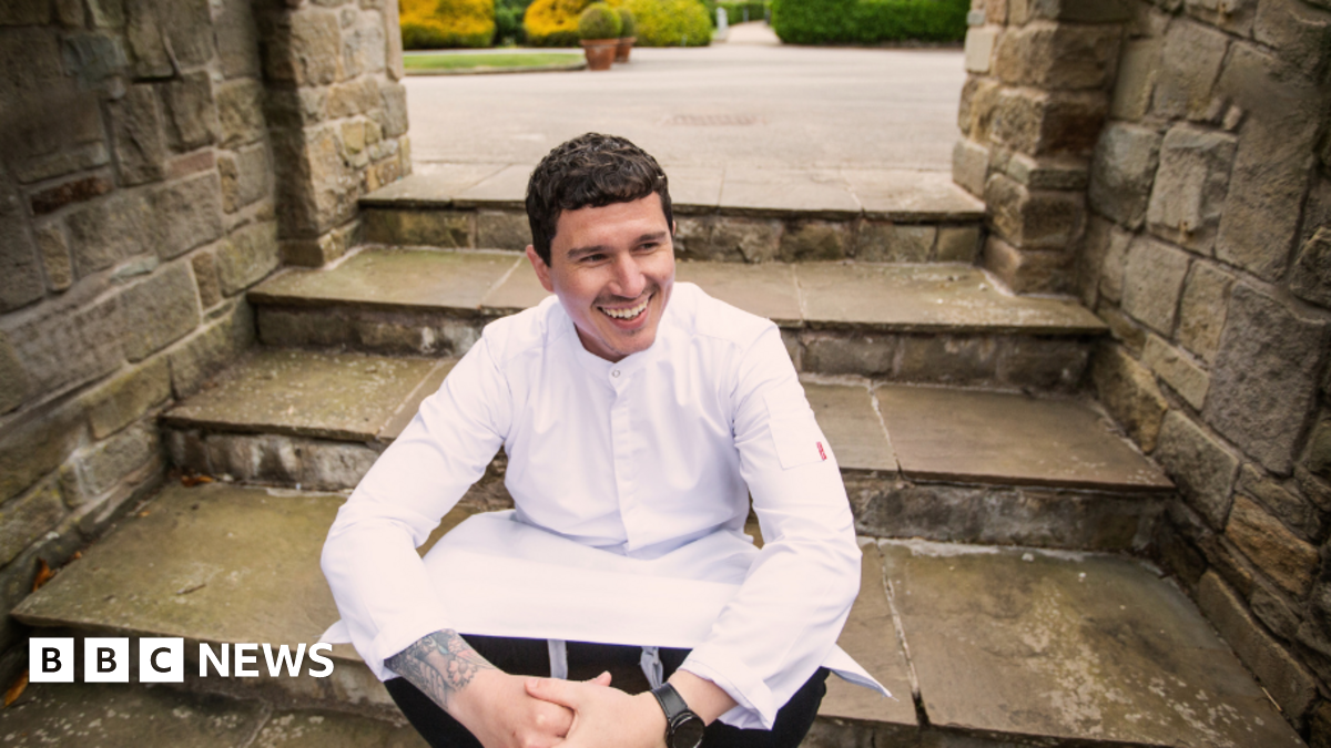 Millie Bobby Brown and David Harbour in character as Eleven and Jim Hopper. Eleven wears red shorts over grey leggings and a grey jumper. Jim Hopper wears a black leather jacket, a black beanie hat and dark trousers.They are speaking to director Frank Darabont on the set of Stranger Things. There is a vintage advertisement posted in the background with debrief on the ground.