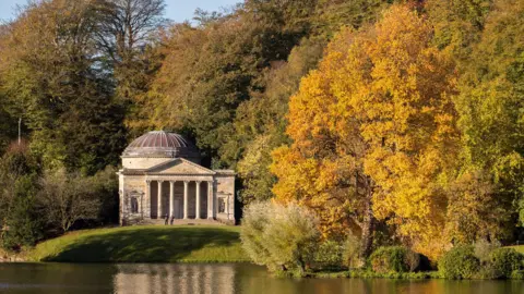 Getty Images Stourhead in Wiltshire. There are orange, green and yellow trees in the shot and a building overlooking water. 