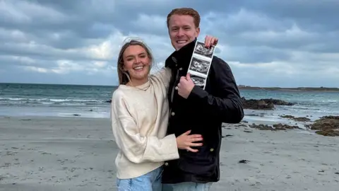 Alastair Chalmers Image shows athlete Alastair Chalmers and his partner on a beach, holding a photo scan of their unborn baby. Chalmers, who has short ginger hair, wears a dark-coloured coat, his partner is pictured in a cream jumper, with jeans and long blond hair. They are both smiling and pictured amidst a backdrop of a choppy sea, rocks and dark grey clouds overhead. 