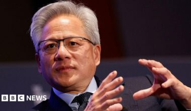 Millie Bobby Brown and David Harbour in character as Eleven and Jim Hopper. Eleven wears red shorts over grey leggings and a grey jumper. Jim Hopper wears a black leather jacket, a black beanie hat and dark trousers.They are speaking to director Frank Darabont on the set of Stranger Things. There is a vintage advertisement posted in the background with debrief on the ground.