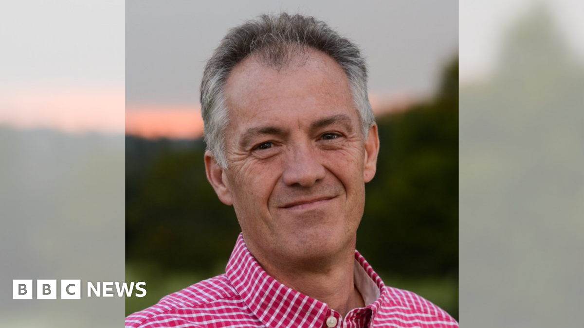 A man with a red and white check shirt smiling at the camera in front of a blurred sunset
