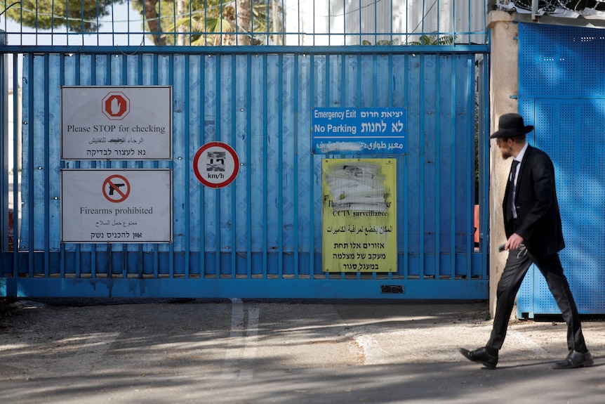 A man in a dark suit and hat walks past a security fence bearing various warning signs.
