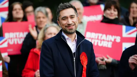 Damian Egan following his election to the newly created seat of Bristol North East during the 2024 general election. He has short dark hair, dark stubble and wears a white shirt, navy jacket and red rosette. A crowd of people behind him hold "Britain's Future" signs. 