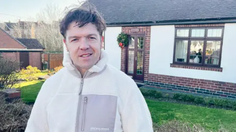 A man with dark hair and a white fleeced jacket with a large grey pocket is standing outside a white house. There is a Christmas wreath beside the door and a small wooden sign above it reads 'Gracelands'. There is a lawn garden with bushes.