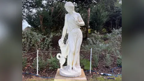 Shuttleworth Trust A close up of the new statue. It is white alabaster. A Greek woman stands looking down at a dog, that is on its hind legs. Its front legs lean against her hips and she pets its head with one hand. Her other arm is angled inwards with her finger wagging at the dog. Her hair is braided back and she wears a loose toga draped across one shoulder and scooped between her legs, so you can see most of her legs. Behind the statue is a roped fence and a shrub border.