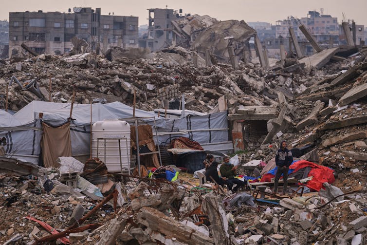 People stand next to a tent set up on top of rubble.