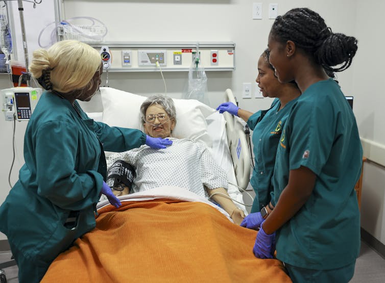 Three young women wearing teal scrubs stand around a dummy of an older woman lying in a hospital bed.