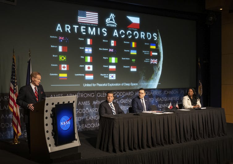 Three people sitting at a panel table and one speaking at a podium with the NASA logo. Projected behind them is a slide reading Artemis Accords, with the flags of several countries.