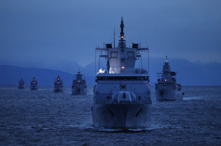 A fleet of military ships at dusk with mountains in the background.
