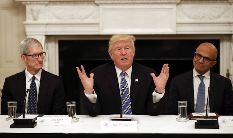 An aged orange-hued man with a gold combover seated between two other men with glum expressions. All wear dark suits.