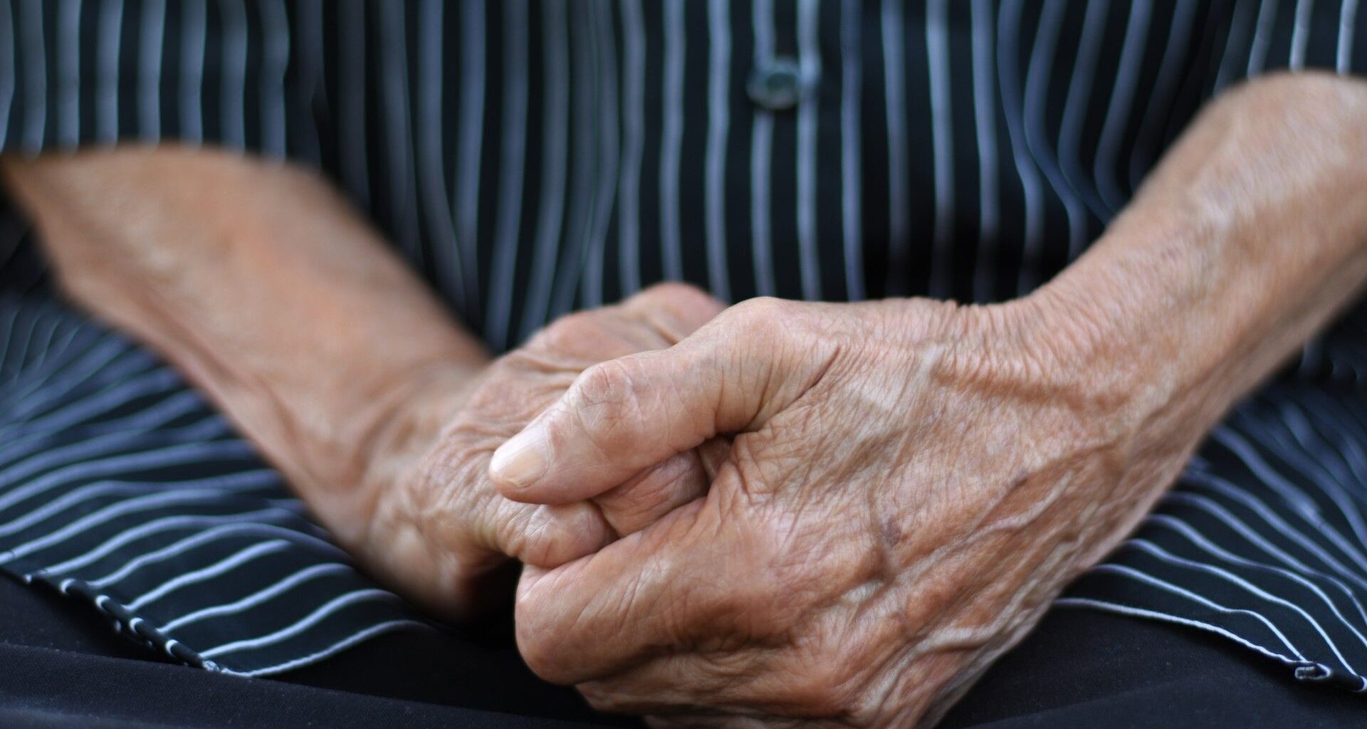 Close-up of the hands of an elderly person that are crossed together over their lap. They are wearing a shirt with black and light blue stripes.
