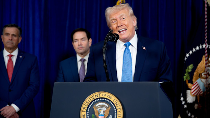 US President Donald Trump speaks at a podium during a news conference, with two unidentified men standing behind him.