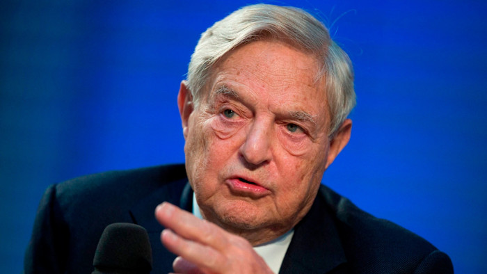 George Soros speaks while gesturing with his hand against a blue background.