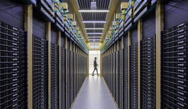 Rows of black server racks line a corridor in the Naver Data Center at GAK Chuncheon, with a person walking in the distance